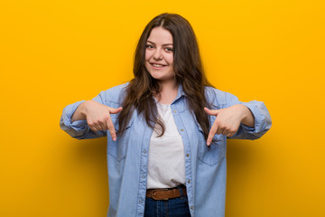 Young curvy plus size woman points down with fingers, positive feeling.