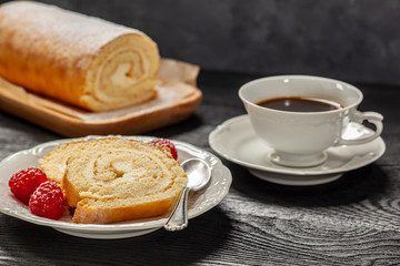 Ciasto - rolada biszkoptowa z kremem mascarpone na talerzyku, obok kawa w filiżance, wszystko postawione na czarnym drewnianym blacie © Grzej