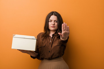 Young curvy plus size woman holding a box standing with outstretched hand showing stop sign,...