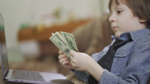 Portrait adorable small boy in jeans jacket sitting on sofa in front of laptop counting the money. A bundle of dollars in child's hands. The leisure of modern kid. Cute son of rich parents