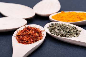 Collection of spices in wooden spoons (Saffron, Basil, Turmeric) on dark background.