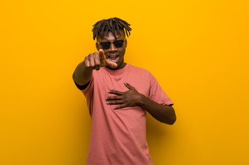 Young casual black man wearing sunglasses points with thumb finger away, laughing and carefree.