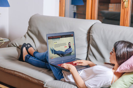 Young woman lying comfortably on the sofa waching video streaming website on laptop. All screen graphics are made up.