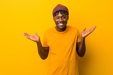 Young black man wearing rastas over yellow background doubting and shrugging him shoulders in...