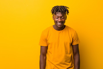 Young black man wearing rastas over yellow background laughs and closes eyes, feels relaxed and...