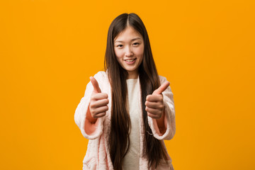 Young chinese woman in pajama with thumbs ups, cheers about something, support and respect concept.