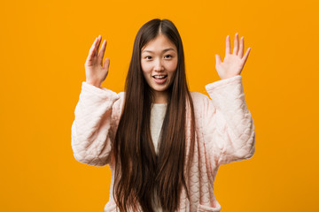 Young chinese woman in pajama receiving a pleasant surprise, excited and raising hands.