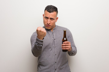Young latin man holding a beer showing fist to front, angry expression