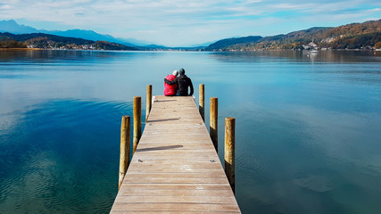 Couple sitting and hugging at the pier by Wörtersee, Pörtschach, Austria. Beautiful lake landscape, surrounded by Alps. This lake is natural drinking water tank. Love is in the air. Romantic moments.