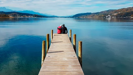 Fotobehang Couple sitting and hugging at the pier by Wörtersee, Pörtschach, Austria. Beautiful lake landscape, surrounded by Alps. This lake is natural drinking water tank. Love is in the air. Romantic moments. © Chris