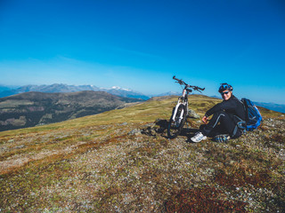 Fototapeta premium A young boy on a mountain bike tour in Millstatt, Austria. Boy wears a big backpack and a helmet. Mountain bike stands next to him. Beautiful mountain view in the back. Professional sport gear.