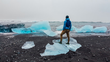 A young man wearing a blue jacket stands on a smaller ice berg, washed to the beach by the sea....