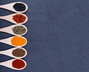 Collection of spices in wooden spoons (Saffron, Basil, Turmeric, Caraway seeds, Sweet Paprika, Black Lava Sea Salt) on dark background.