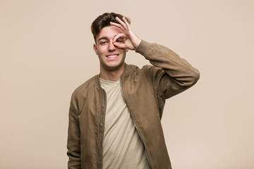 Young caucasian man wearing a brown jacket excited keeping ok gesture on eye.