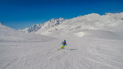 Naklejka na ściany i meble A skier going down the slope in Heiligenblut, Austria. Perfectly groomed slopes. High mountains surrounding the man wearing yellow trousers and blue jacket. Man wears helm for the protection.