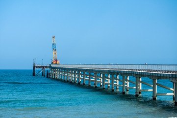 Fototapeta na wymiar View to pier with industrial crane among calm sea waters on blue sky background in clear summer day