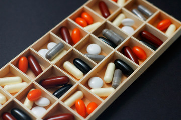 Set of different pills and capsules in wooden pillbox for daily and week dosage. On black background. Hospital medicine box.