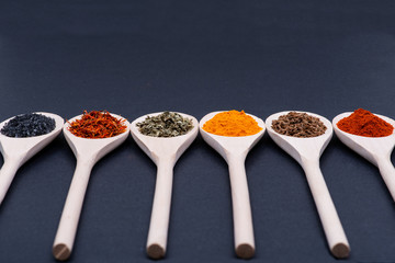 Collection of spices in wooden spoons (Saffron, Basil, Turmeric, Caraway seeds, Sweet Paprika, Black Lava Sea Salt) on dark background.