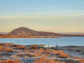 A flatland, covered with golden grass. Dried grass due to the winter season. Fjord view, with some taller mountains in the back. Nice and soft colors of the sunrise.