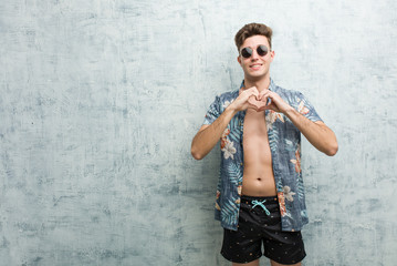 Young caucasian man wearing a swimsuit smiling and showing a heart shape with him hands.