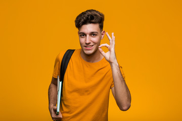 Young student man holding books surprised pointing at himself, smiling broadly.