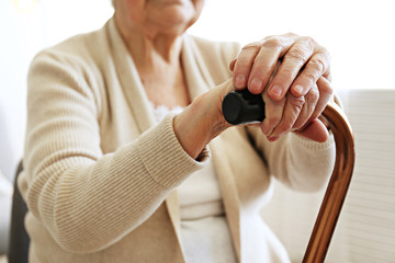 Elderly woman sitting in nursing home room holding walking quad cane with wrinked hand. Old age senior lady wearing beige cardigan, metal aid stick handle bar close up. Interior background, copy space