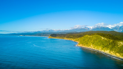 Obraz premium Remote rocky coastline with native forest and snowy mountain peaks on the background. West Coast, South Island, New Zealand