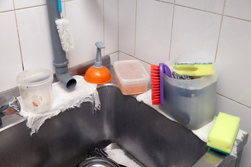 Close-up of a metal sink in the professional kitchen of the restaurant, orange plunger, pipe, faucet, container with dishwashing liquid, sponges, brushes, rags, washcloths
