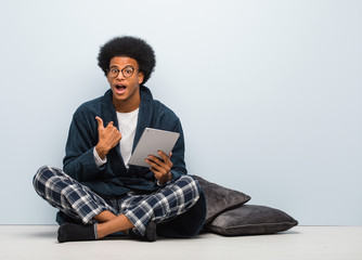 Young black man sitting on his house and holding his tablet surprised, feels successful and prosperous