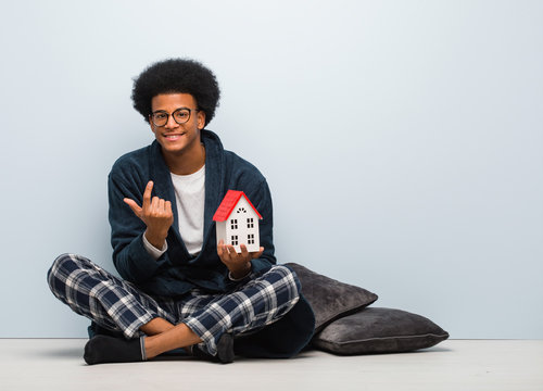 Young black man holding a house model sitting on the floor inviting to come