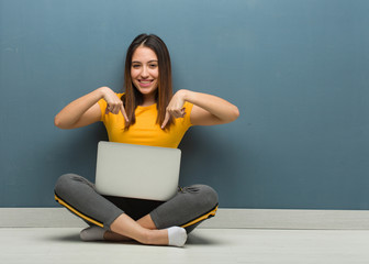 Young woman sitting on the floor with a laptop pointing to the bottom with fingers