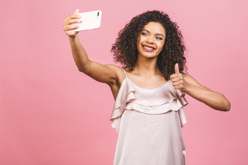 Portrait of a pretty young afro american woman in dress while standing and taking a selfie isolated over pink background.