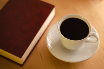 Close view of a full coffee cup with a book next to it, resting on a wooden table, illuminated by a soft and warm light