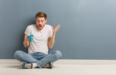 Young redhead student man sitting on the floor celebrating a victory or success. He is holding a coffee mug.