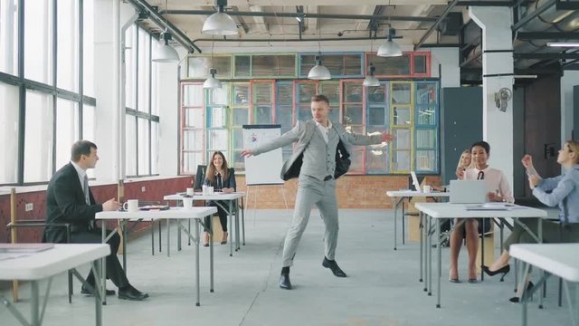 A young businessman in a business suit enters the office, starts to dance expressively, grabs a glass from the employee's table and throws it. Coworking in loft style. Office life. Celebration