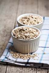 Raw oat flakes in ceramic bowls on linen napkin, golden wheat ears on rustic wooden background.