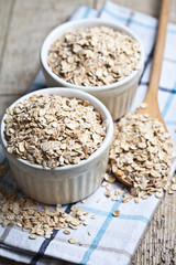 Oat flakes in ceramic bowls and wooden spoon on linen napkin, golden wheat ears on rustic wooden background.