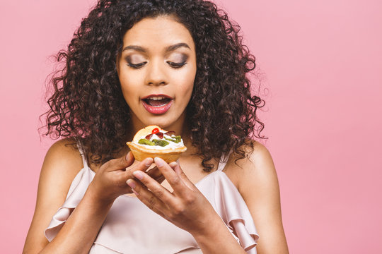 Black American African Happy Woman With Curly Afro Hair Style Making A Mess Eating A Huge Fancy Dessert Over Pink Background. Eating Cupcake.