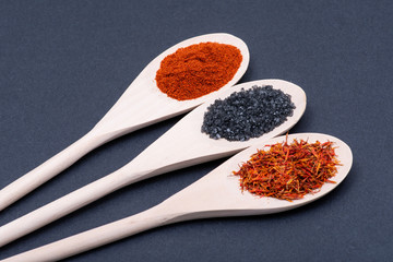 Saffron, Black Lava Sea Salt and Sweet Paprika in wooden spoons on dark background.