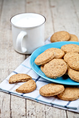 Fresh baked oat cookies on blue ceramic plate on linen napkin and cup of milk on rustic wooden table.