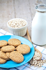 Fresh baked oat cookies on blue ceramic plate on linen napkin, bottle of milk and oak flakes on rustic wooden table.