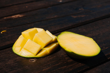 Two slices of a ripe mango one with cubes placed on a wooden surface during day time – Bright yellow exotic juicy fruit on brown background – Healthy and delicious snack