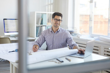 Portrait of smiling engineer working with plans and blueprints sitting at desk in office, copy space