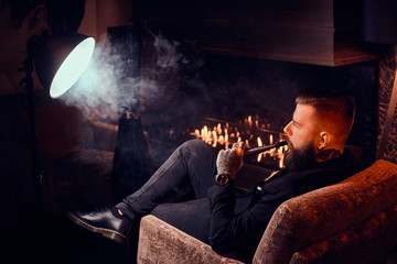 Bearded handsome man is sitting near fireplace and smoking hookah, making good misty vapour.