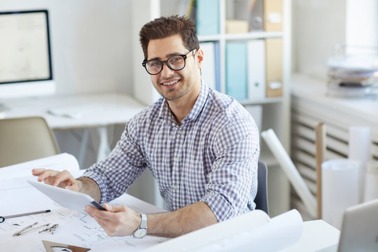 Portrait Of Young Engineer Smiling At Camera While Using Digital Tablet Sitting At Desk In Office, Copy Space