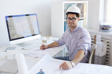 Portrait of contemporary engineer wearing hardhat sitting at desk in office, copy space