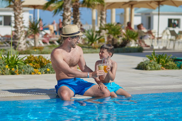 Dad and son enjoying their summer holidays. They are holding their cocktails in the hands, while sitting near the hotel’s swimming pool.