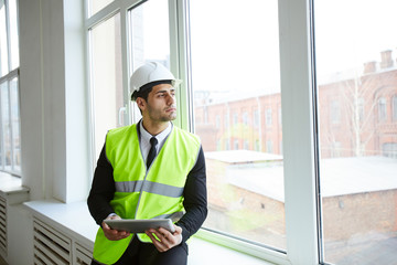 Waist up portrait of handsome Middle-Eastern businessman wearing hardhat looking at window at construction site, copy space
