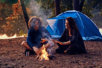Attractive romantic couple is warming up near bonfire in the evening forest. There are tent and river nearby.