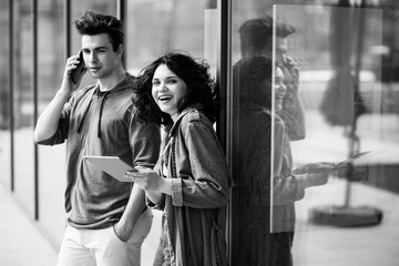 Young cute couple - a boy and a girl standing near a glass building. The couple spend time together. The guy speaks on the phone, and the girl holds a tablet in her hands